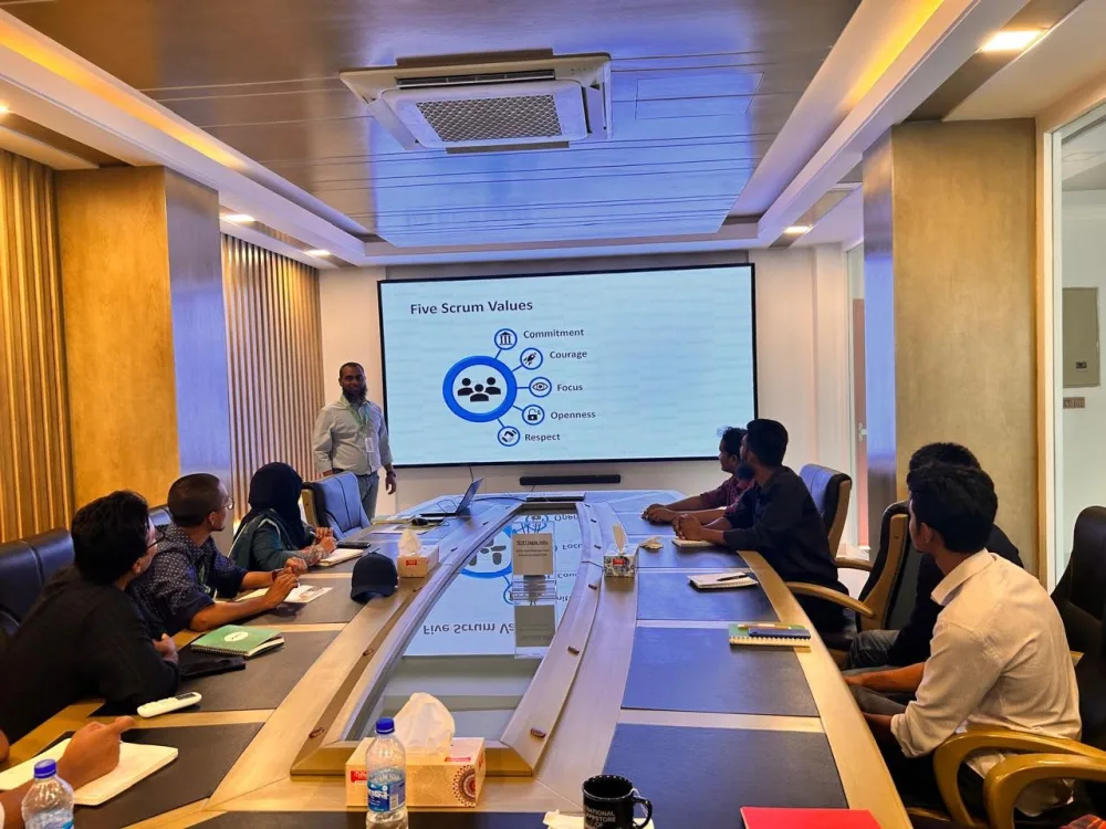A trainer beside a large screen showing the five Scrum values, with a small group taking notes around a boardroom table